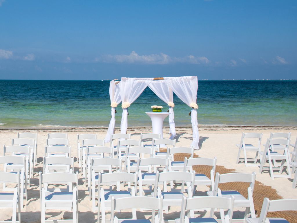 Wedding ceremony with a large gathering on the beach, featuring a draped arch facing the sea.