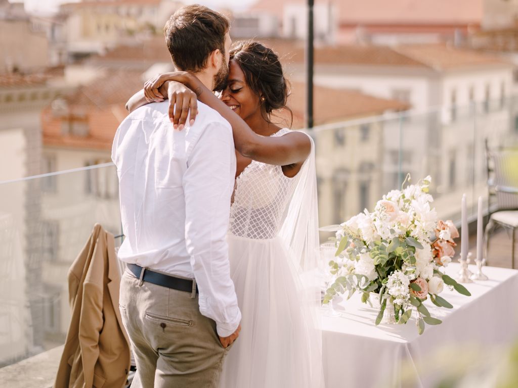 Young newlyweds embracing on a balcony with panoramic view, intimate seaside wedding in the Caribbean with white flower bouquet and greenery