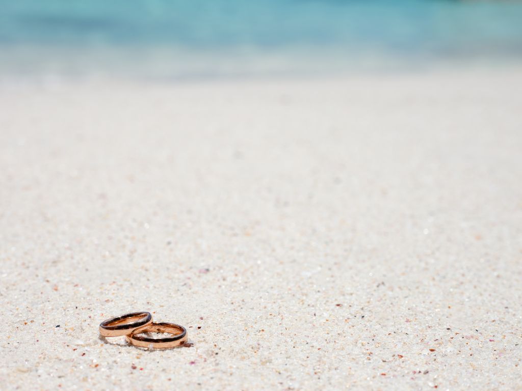 Wedding rings on beach sand with the sea in the background, symbolizing a Caribbean wedding.